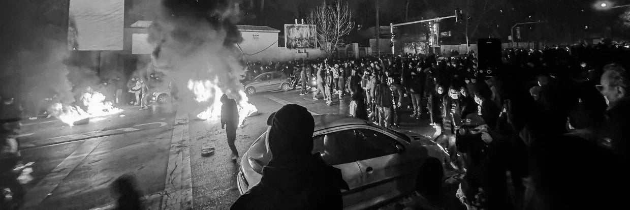 Night protest scene with crowd, burning barricades, and cars under streetlights