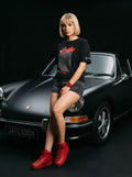 Woman in black Persian graphic tee, denim shorts, red sneakers, posing by classic black car