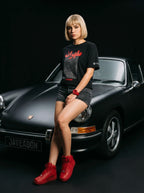 Woman in black Persian graphic tee, denim shorts, red sneakers, posing by classic black car