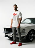 Man in white Tehran graphic tee with red sneakers, standing beside classic white muscle car