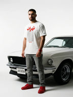 Man in white Tehran graphic tee with red sneakers, standing beside classic white muscle car