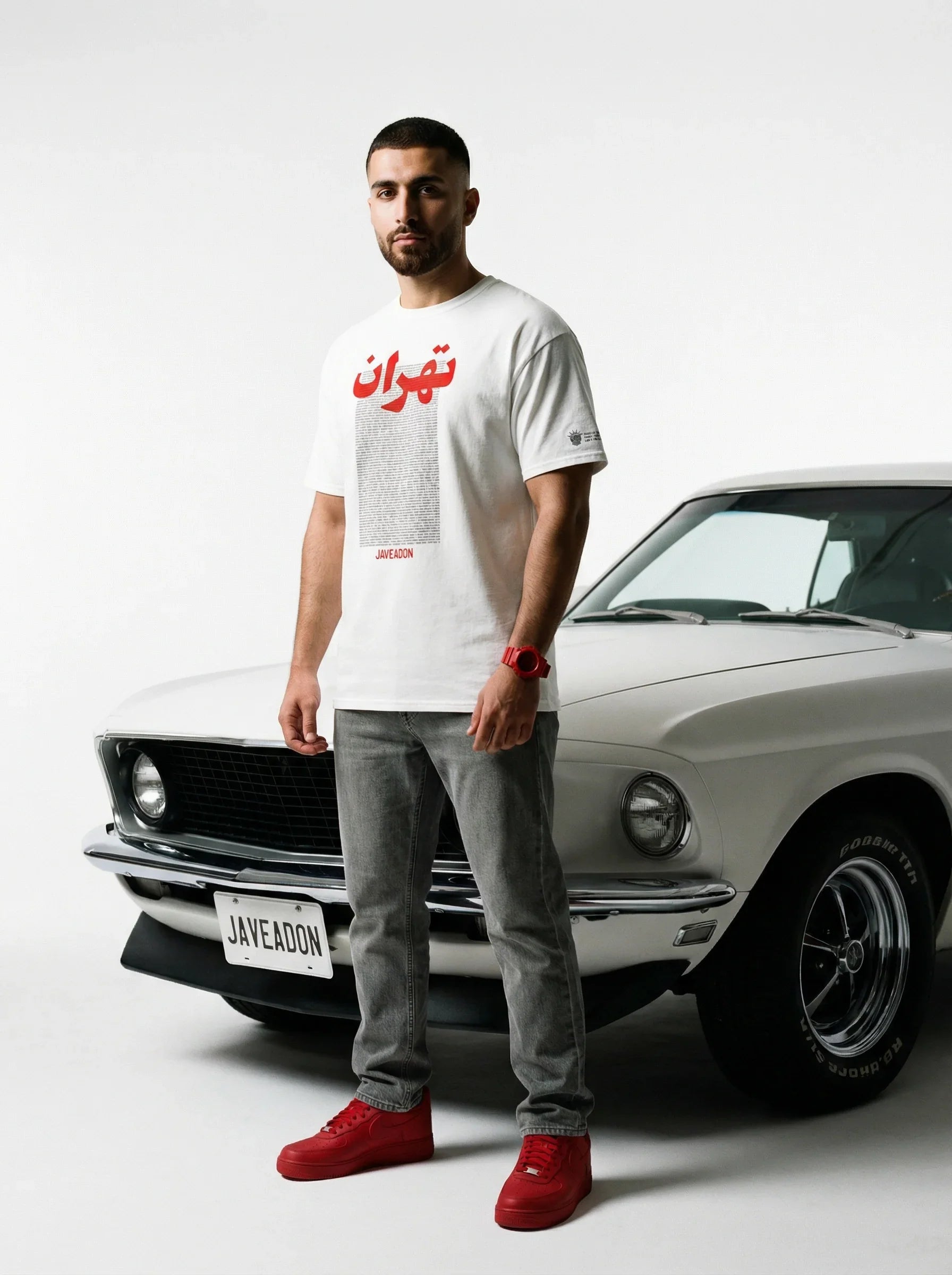 Man in white Tehran graphic tee with red sneakers, standing beside classic white muscle car