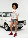 Woman in white Persian-themed graphic tee, gray shorts, red sneakers, standing by vintage white car