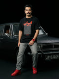 Man wearing black Persia-themed streetwear tee with red accents, gray jeans, red sneakers, posing by vintage car