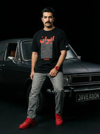 Man wearing black Persia-themed streetwear tee with red accents, gray jeans, red sneakers, posing by vintage car