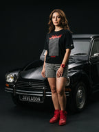 Woman in black Javeadon t-shirt with Persian script, red sneakers, standing beside classic black car