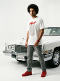 Man in white Persian-themed graphic tee, grey jeans, red sneakers, standing by classic white car