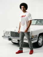 Man in white Persian-themed graphic tee, grey jeans, red sneakers, standing by classic white car