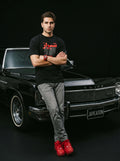 Man in black Persian-themed tee and red sneakers leaning on classic black Buick, embodying modern streetwear style.