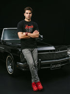 Man in black Persian-themed tee and red sneakers leaning on classic black Buick, embodying modern streetwear style.