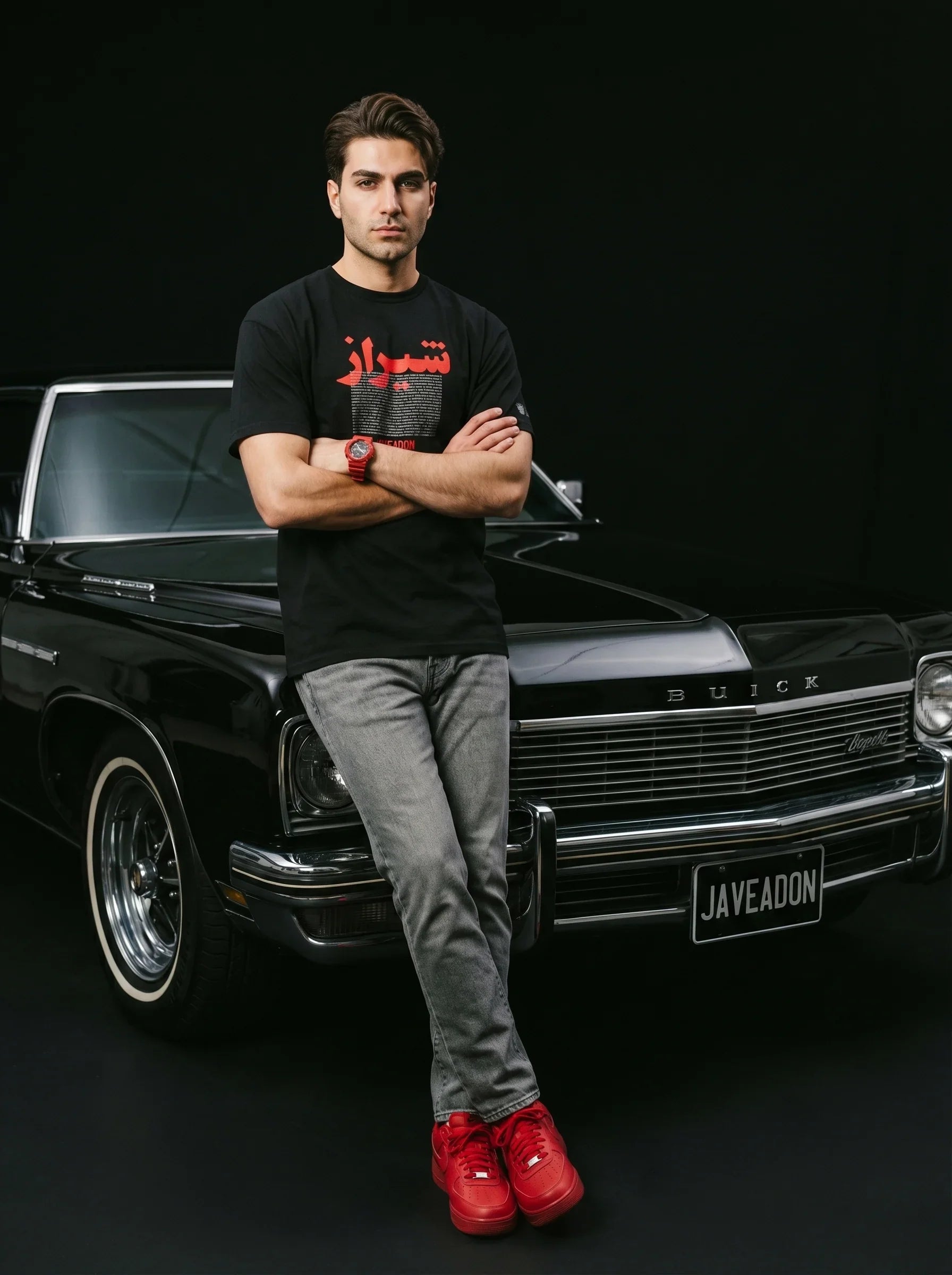 Man in black Persian-themed tee and red sneakers leaning on classic black Buick, embodying modern streetwear style.