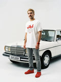 Man in white Persian graphic tee and red sneakers stands beside classic white Mercedes, showcasing streetwear style.