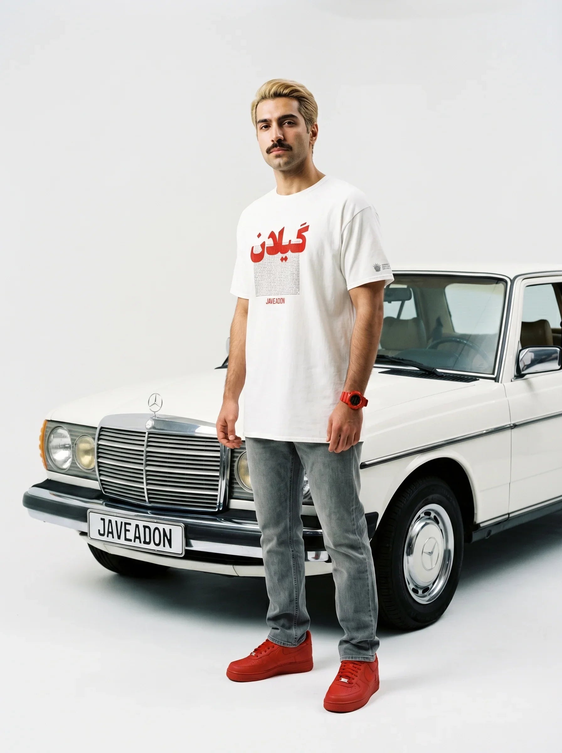 Man in white Persian graphic tee and red sneakers stands beside classic white Mercedes, showcasing streetwear style.