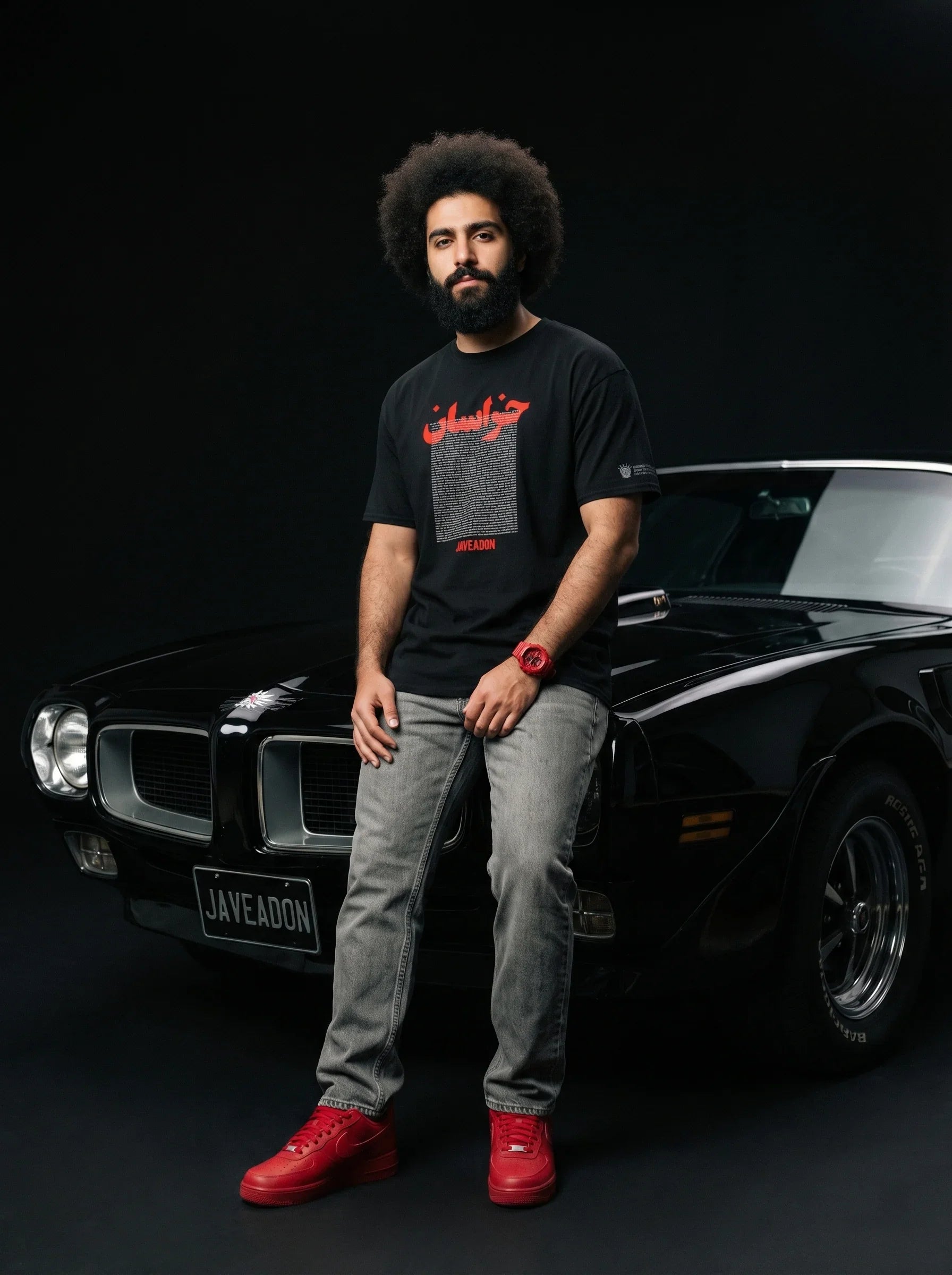Man in black Persian-inspired streetwear tee with red accents, leaning on classic black muscle car, bold red sneakers