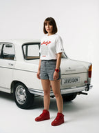 Woman in white Persian-themed tee, denim shorts, red sneakers, standing by vintage car