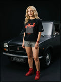 Woman in black Persian graphic tee, denim shorts, red sneakers, standing by classic black Fiat car