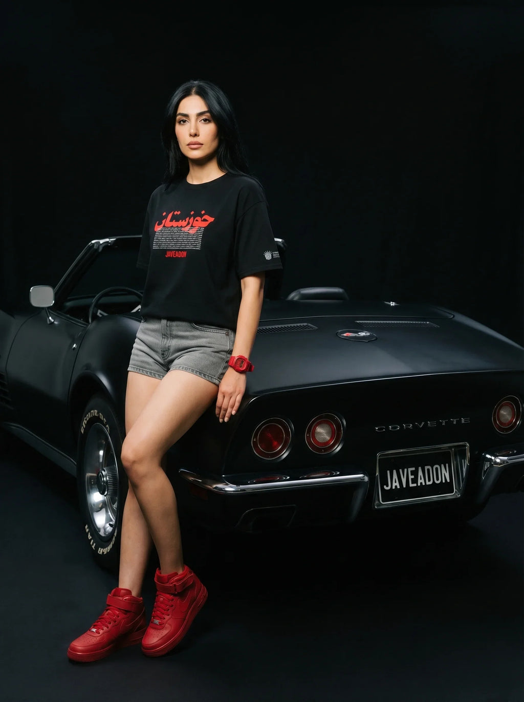 Woman in black Persian graphic tee and red sneakers leaning on classic black Corvette, bold streetwear style