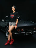 Woman in black Persian graphic tee and red sneakers leaning on classic black Corvette, bold streetwear style