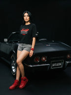 Woman in black Persian graphic tee and red sneakers leaning on classic black Corvette, bold streetwear style