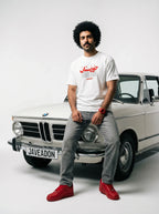 Man in white Persian graphic tee and red sneakers leaning on vintage BMW, streetwear style