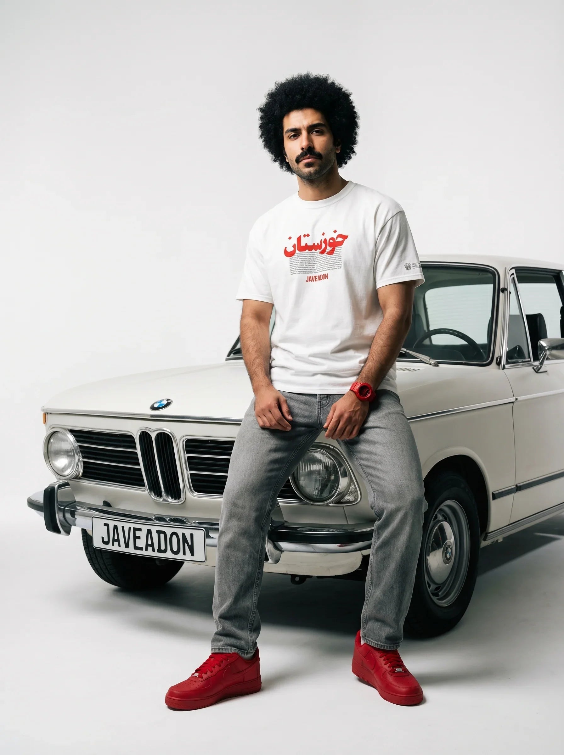 Man in white Persian graphic tee and red sneakers leaning on vintage BMW, streetwear style