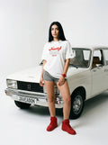 Woman in white Persian-themed tee and red sneakers posing beside vintage car with Javeadon license plate