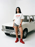 Woman in white Persian-themed tee and red sneakers posing beside vintage car with Javeadon license plate