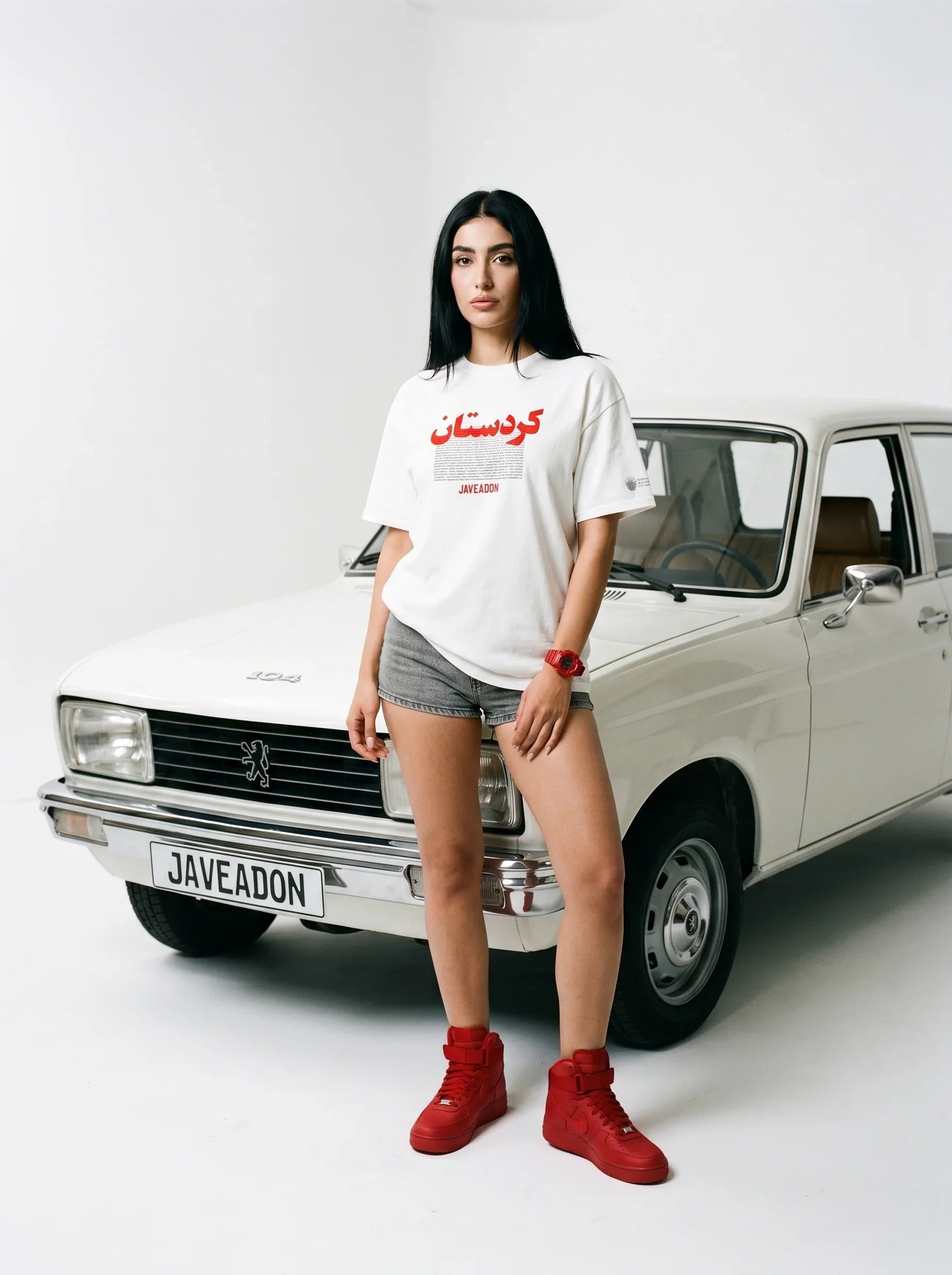Woman in white Persian-themed tee and red sneakers posing beside vintage car with Javeadon license plate
