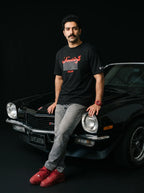 Man in black Persian graphic tee and red sneakers leaning on classic black muscle car, bold streetwear style