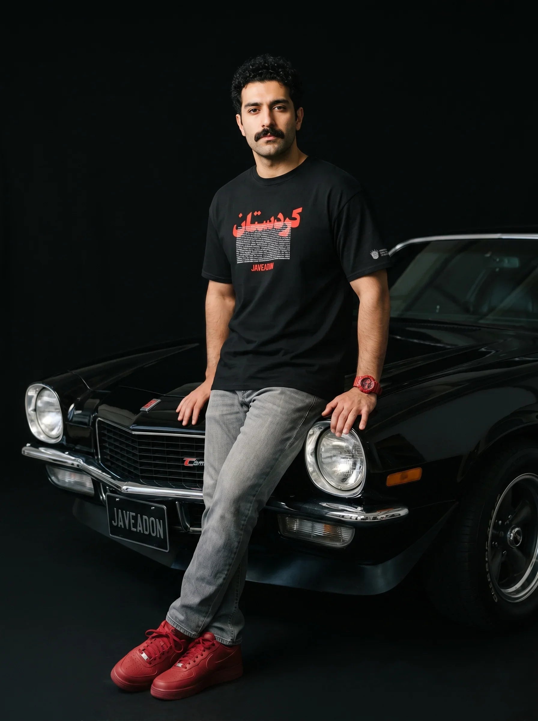 Man in black Persian graphic tee and red sneakers leaning on classic black muscle car, bold streetwear style