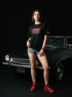 Woman in black Persian graphic tee and red sneakers poses by classic Ford Pinto, bold streetwear style