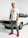 Man in white Persian graphic tee, grey jeans, red sneakers, standing by classic white muscle car