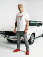 Man in white Persian graphic tee, grey jeans, red sneakers, standing by classic white muscle car