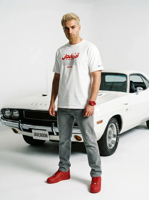 Man in white Persian graphic tee, grey jeans, red sneakers, standing by classic white muscle car