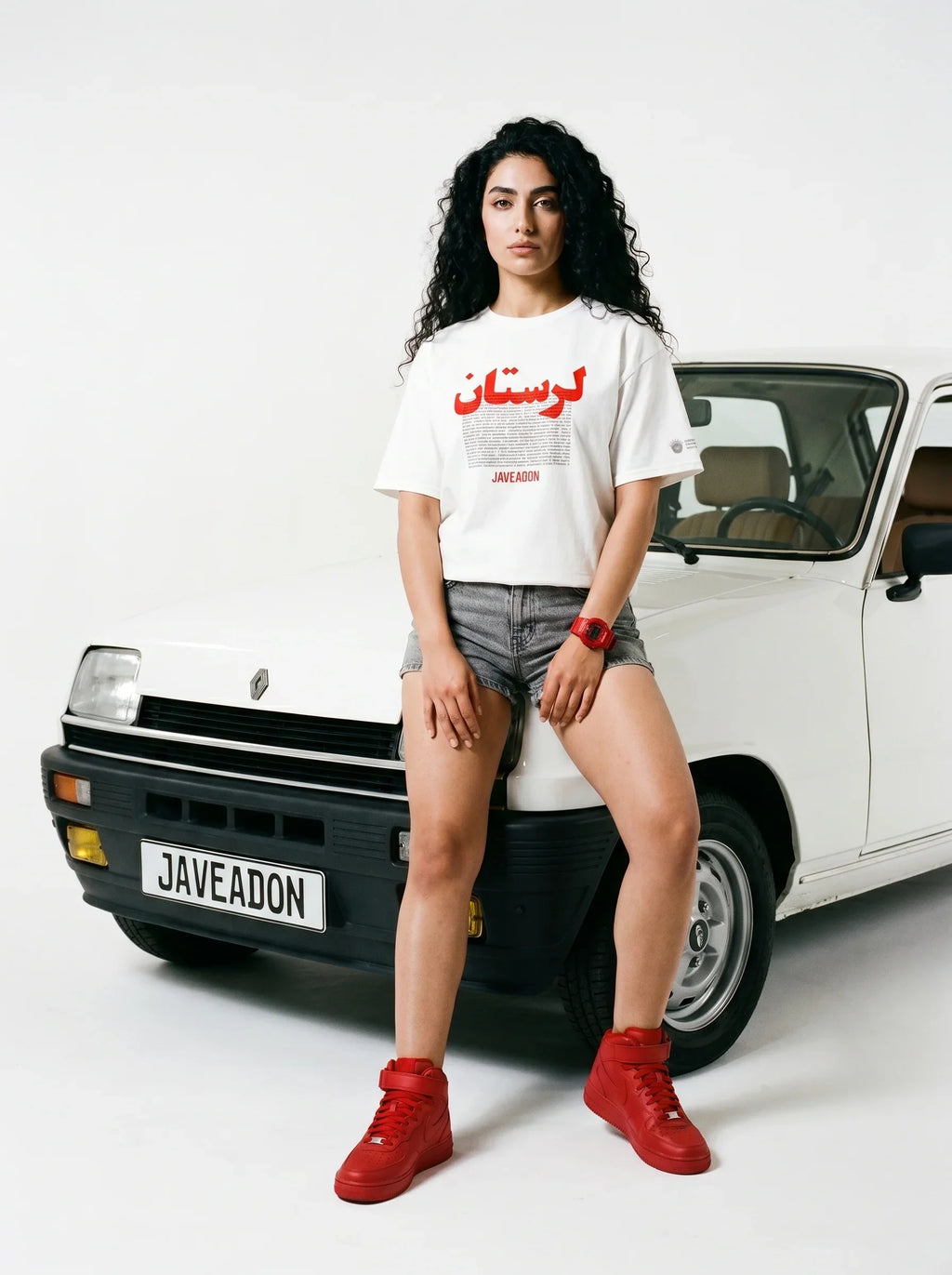 Woman in white Persian-themed tee, denim shorts, red sneakers, leaning on vintage car with Javeadon license plate