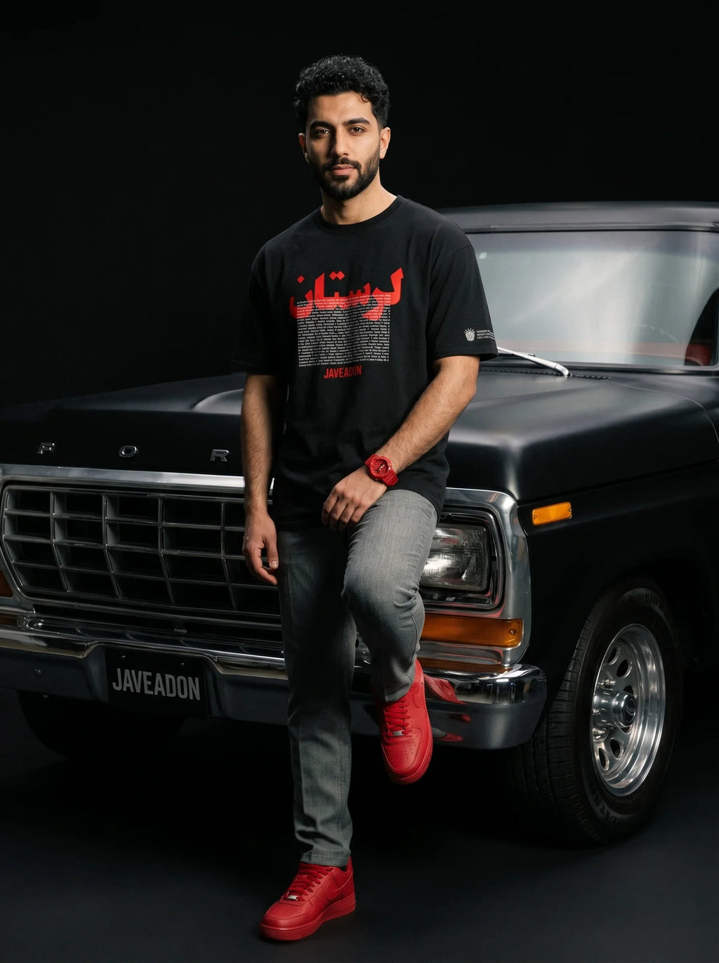 Man in black Persian streetwear tee with red graphics, leaning on classic Ford truck, bold red sneakers