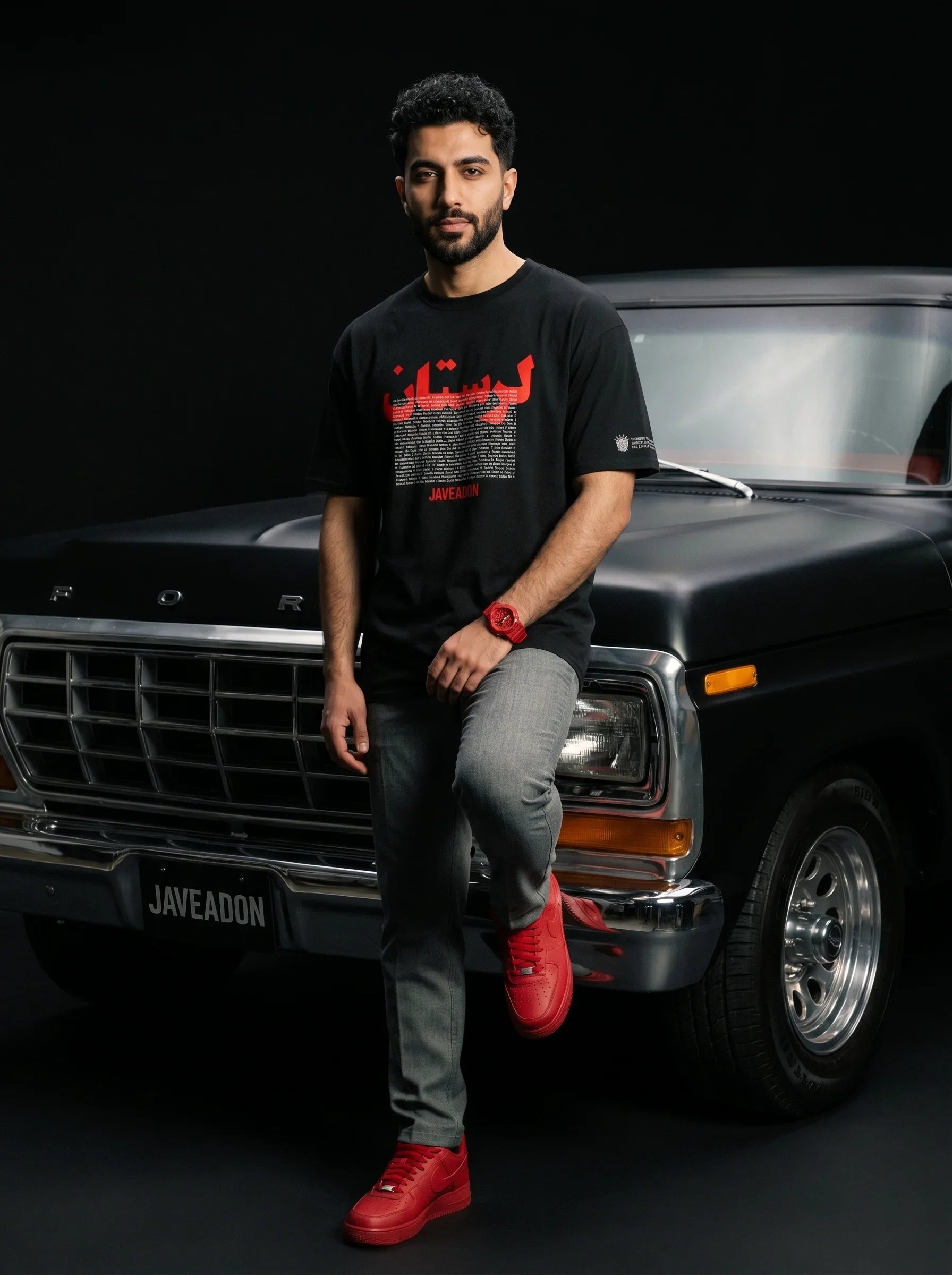 Man in black Persian streetwear tee with red graphics, leaning on classic Ford truck, bold red sneakers