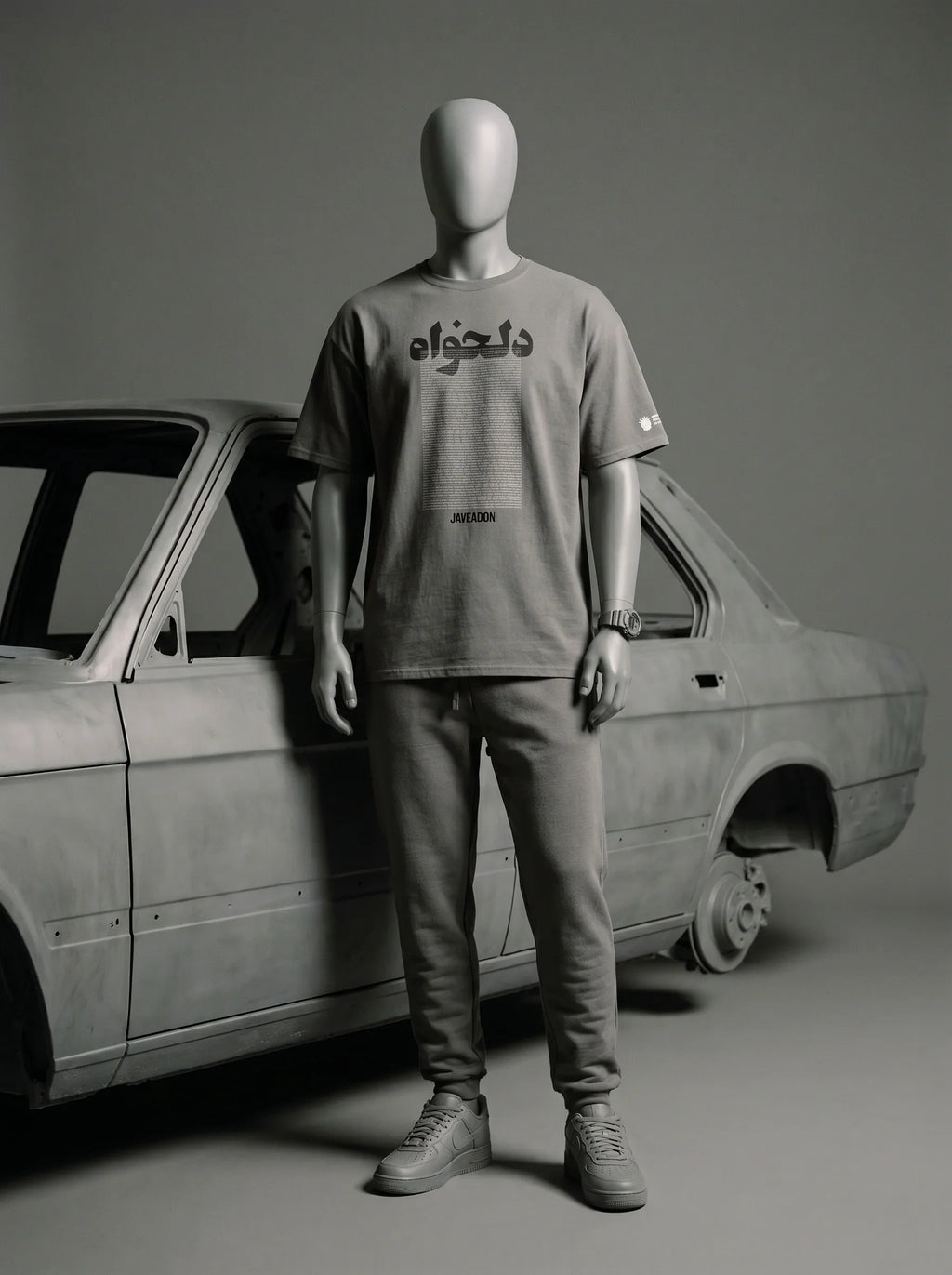Mannequin wearing gray streetwear tee with Persian text, paired with joggers, beside vintage car.