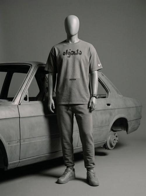 Mannequin wearing gray streetwear tee with Persian text, paired with joggers, beside vintage car.