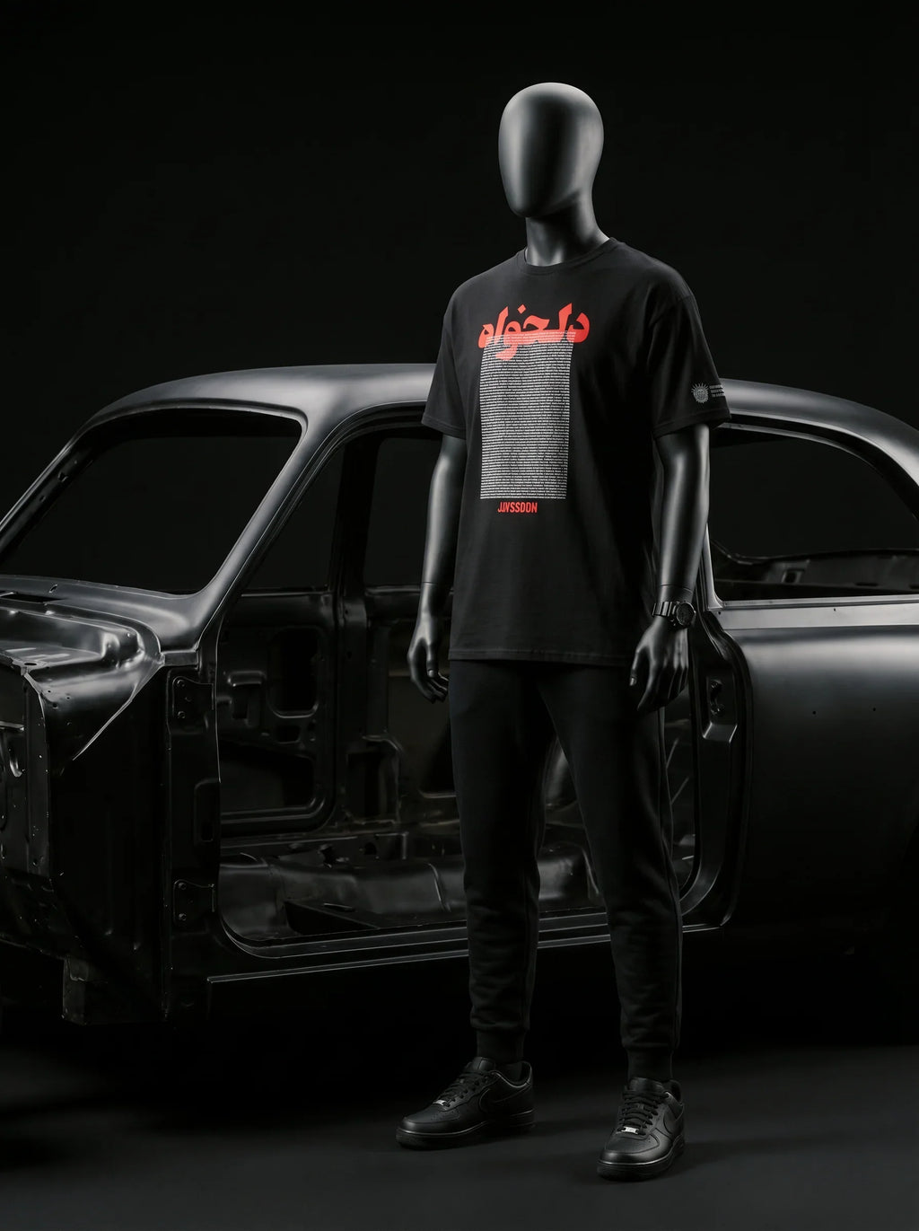 Black mannequin wearing Persian-themed streetwear tee with red text, styled with black joggers and sneakers near car frame