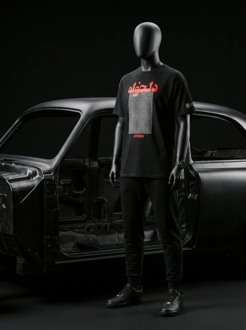 Black mannequin wearing Persian-themed streetwear tee with red text, styled with black joggers and sneakers near car frame