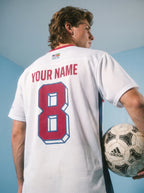 Custom white soccer jersey with red number 8 and 'YOUR NAME', held with Adidas ball, featuring heritage flag patch.